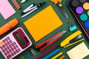 An overhead view of tools used in an office or to make art - a calculator, pad of paper, boxcutters, protractor, pencils, scissors.